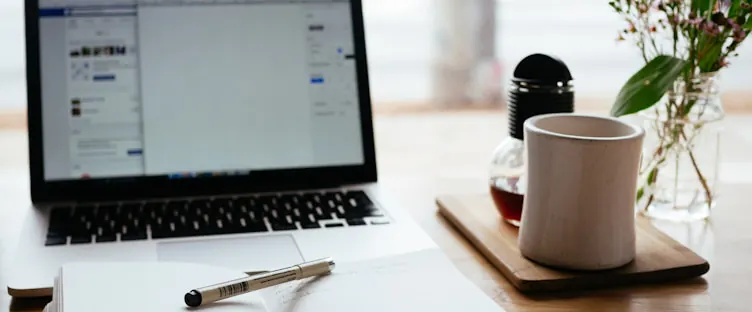 An open laptop on a desk beside a notebook, pen, and a cup of tea