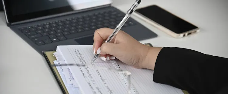 A hand writing in a notebook beside a keyboard and a phone