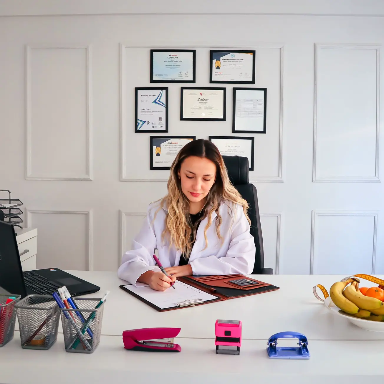 A nutritionist sitting at her desk writing on a clipboard, a laptop to one side and a bowl of fruit to the other