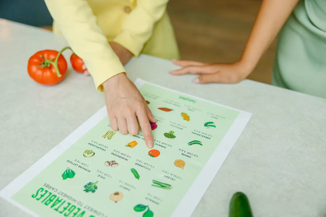 A nutritionist's hand pointing to an item on a printed vegetable-portion chart laid on a table, fresh tomatoes beside it and the client's hands resting opposite