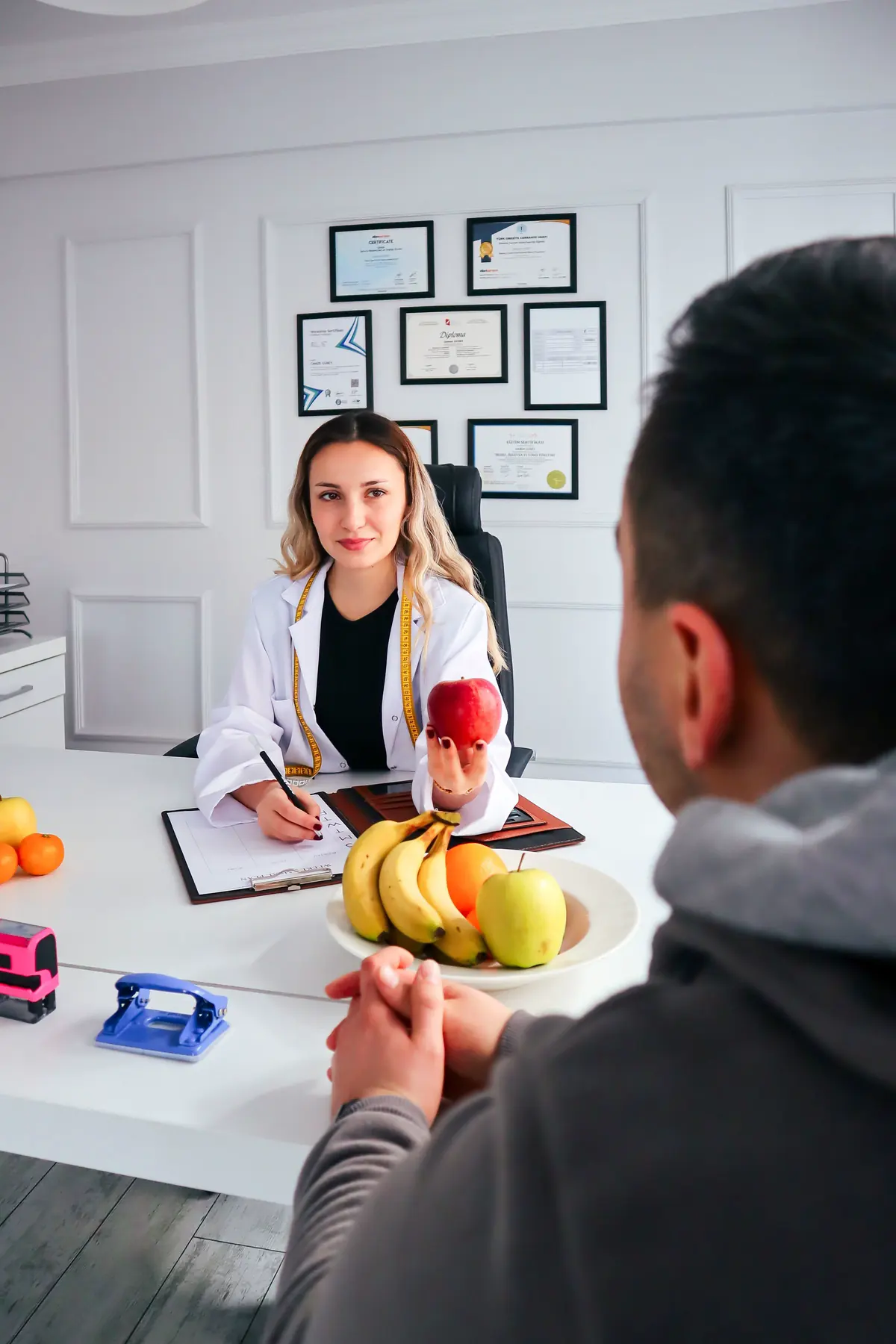 A nutritionist in a white coat at her desk, a tape measure round her neck, holding out an apple to a client whose back is to the camera, framed qualifications on the wall behind her