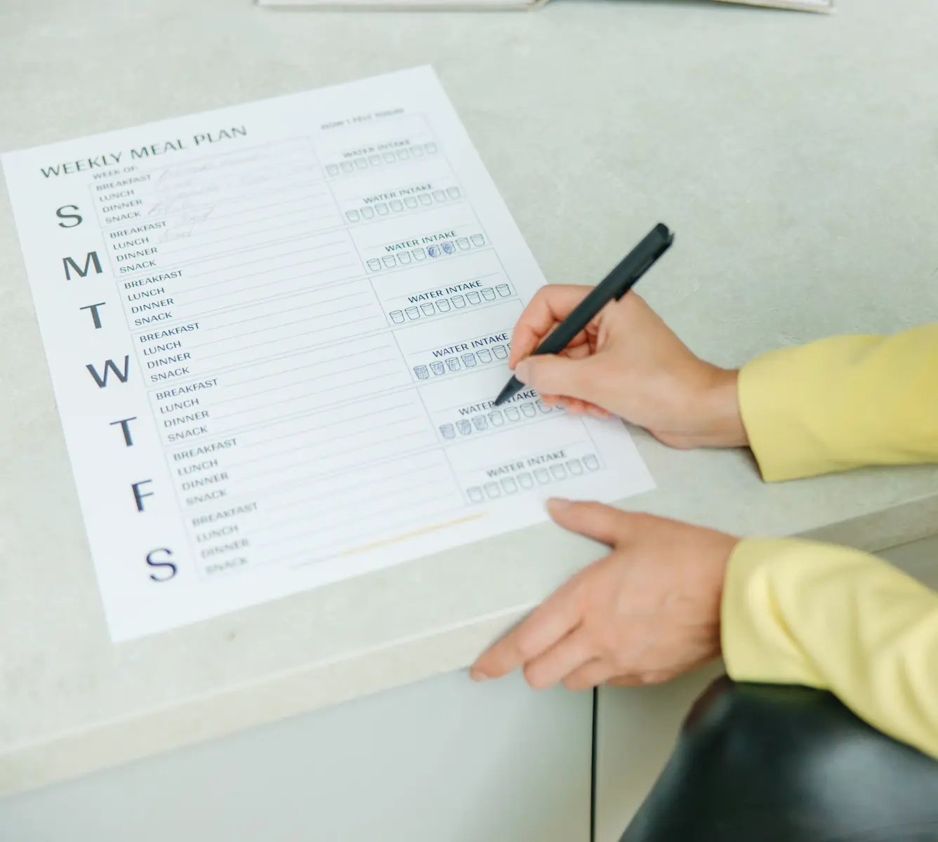 A hand filling in a printed weekly meal plan by hand, the days of the week down the left edge and every meal slot still blank