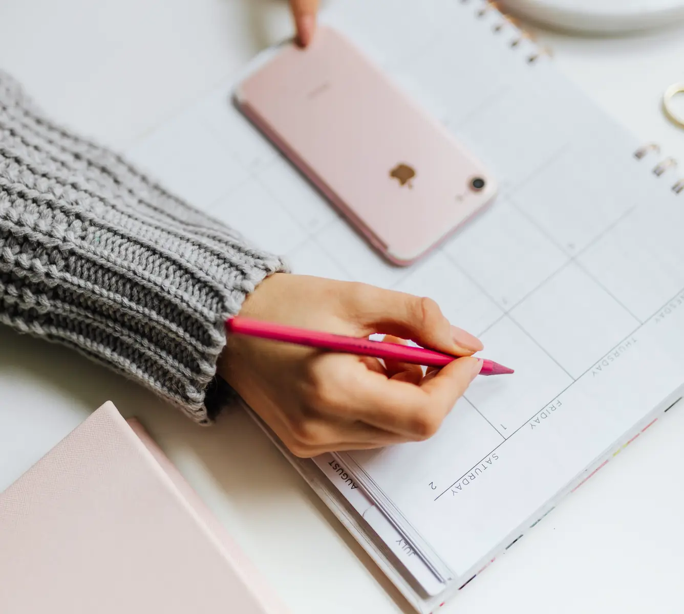 A hand marking a date in a paper month planner, a phone lying face down across the empty week above it