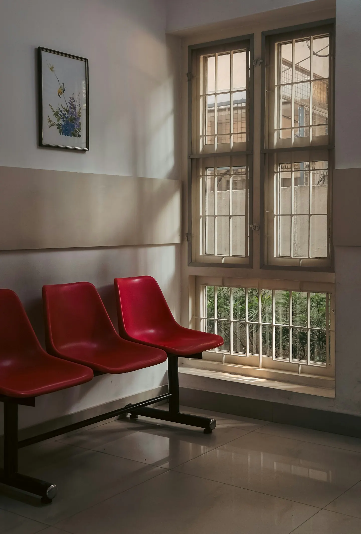 An empty waiting area, red seats beside a tall barred window in afternoon light